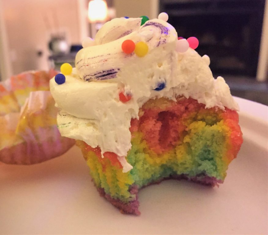 Photo of a rainbow cupcake with half of it bitten off, so you can see the colors inside the mix.