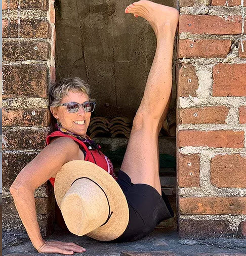 Image of Beth Goldstein wearing shorts, sunglasses with a straw hat by her side, doing a yoga pose.